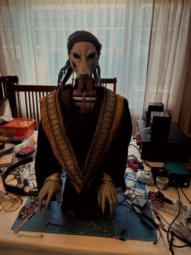 Grandmother Lupa dressed: headscarf, embroidered shawl and clawed hands resting on the cutting mat