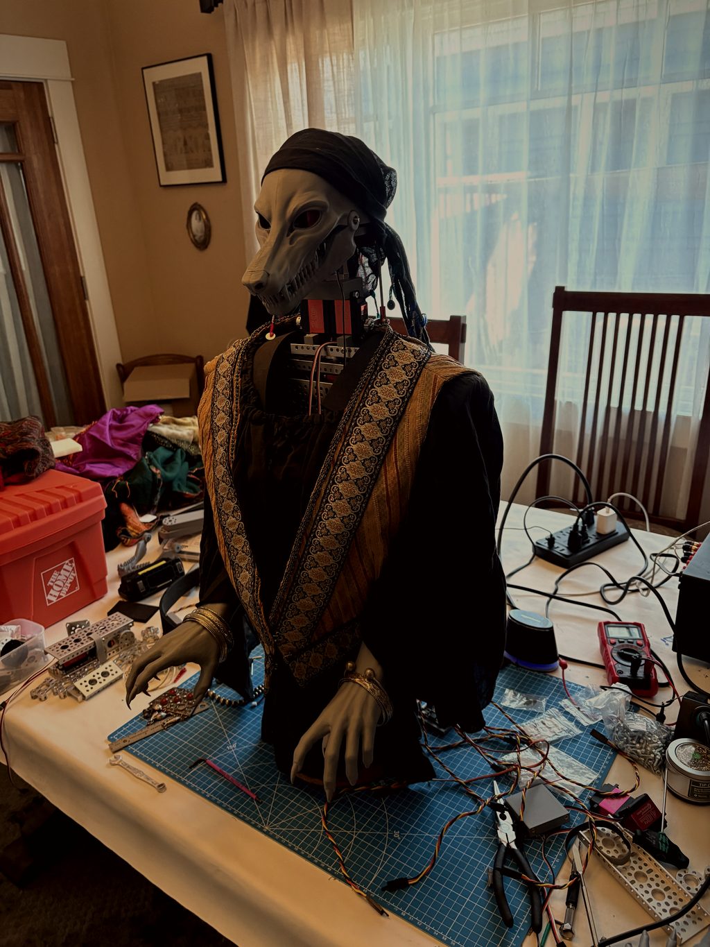 Grandmother Lupa at the workbench in her headscarf and embroidered shawl, clawed hands resting on the table