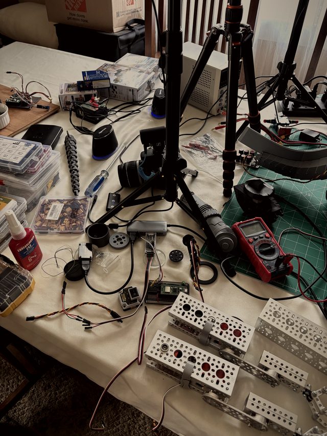Electronics workbench with camera tripods, multimeter, and aluminum servo brackets wired with servo leads