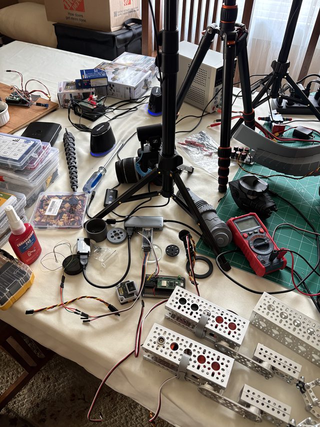 Electronics workbench with camera tripods, multimeter, and aluminum servo brackets wired with servo leads